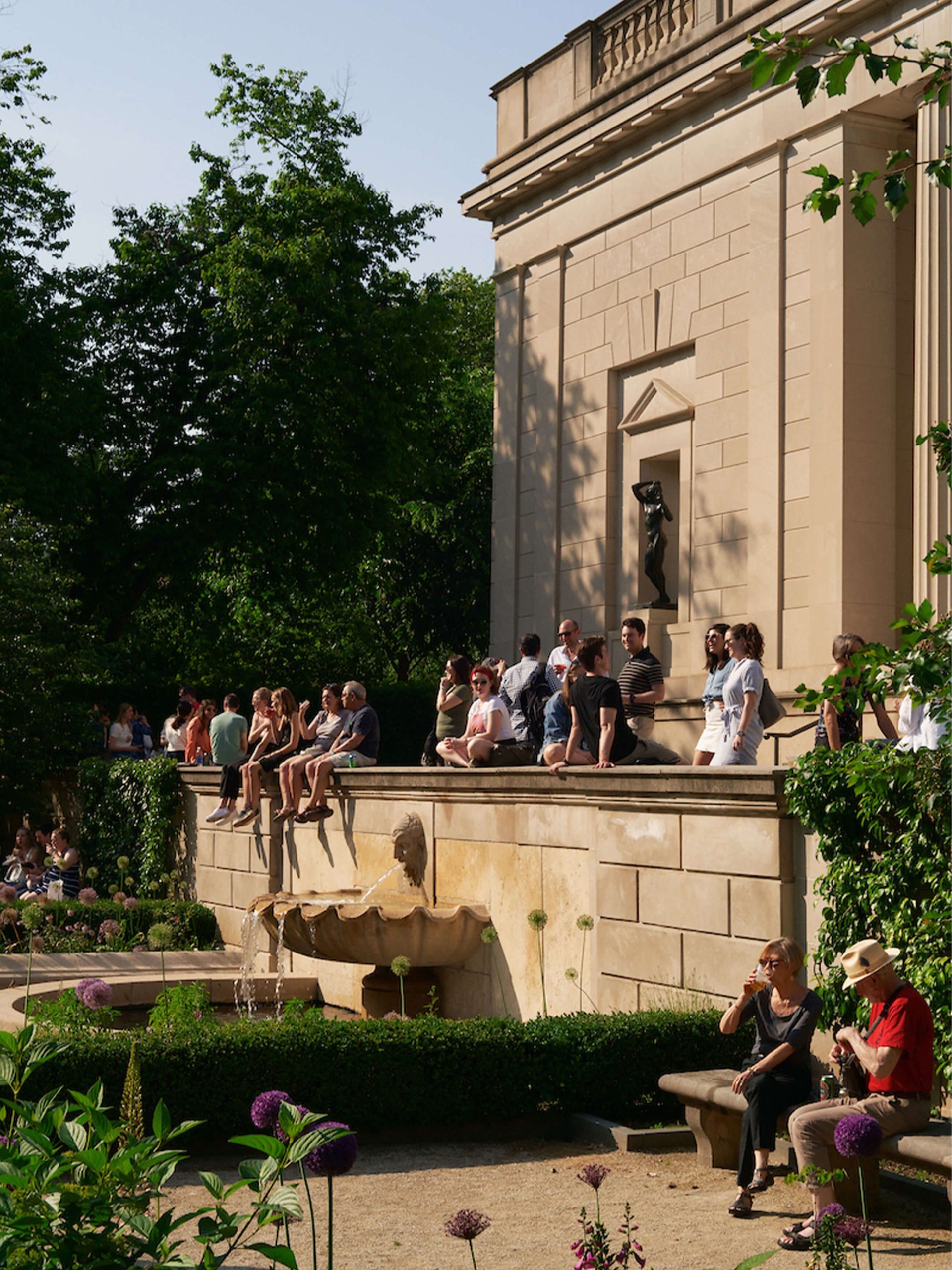 The Rodin Museum Garden Bar Returns May 29 With a New Food Truck and a Crossing Choir Performance 3 Visitors gather at golden hour on the stone wall of the Rodin Museum during a summer Garden Bar evening with a bronze Rodin sculpture visible in the portico niche.