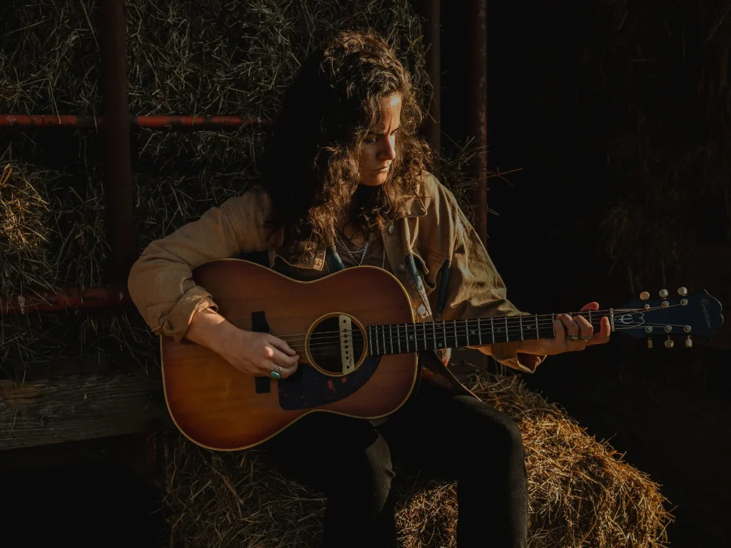 Alyssa Hankey, a Pittsburgh-based Americana singer-songwriter, sits with an acoustic guitar on hay bales in a barn setting