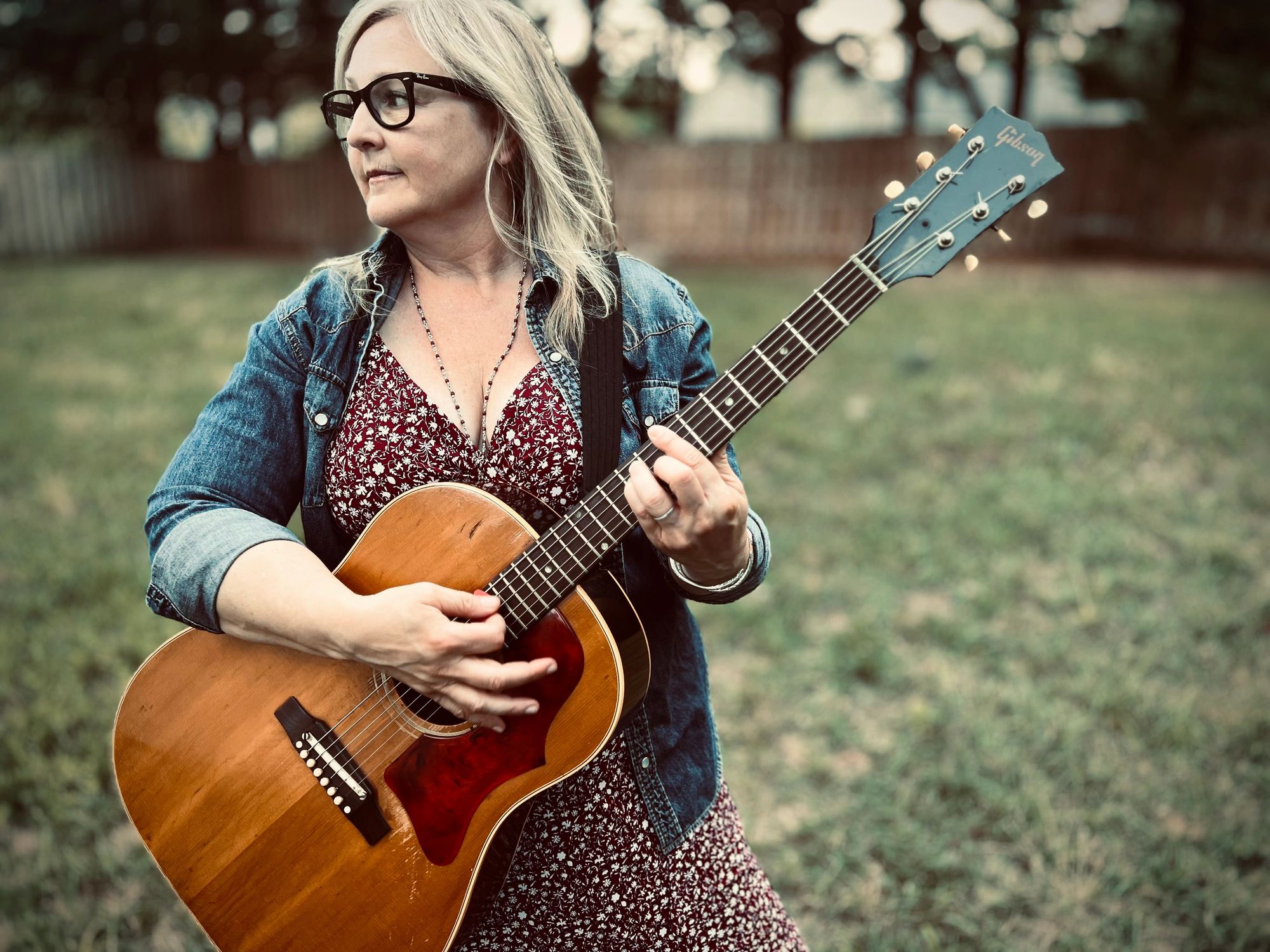 Meg Murray, an Annapolis singer-songwriter, stands outdoors in a denim jacket holding an acoustic guitar