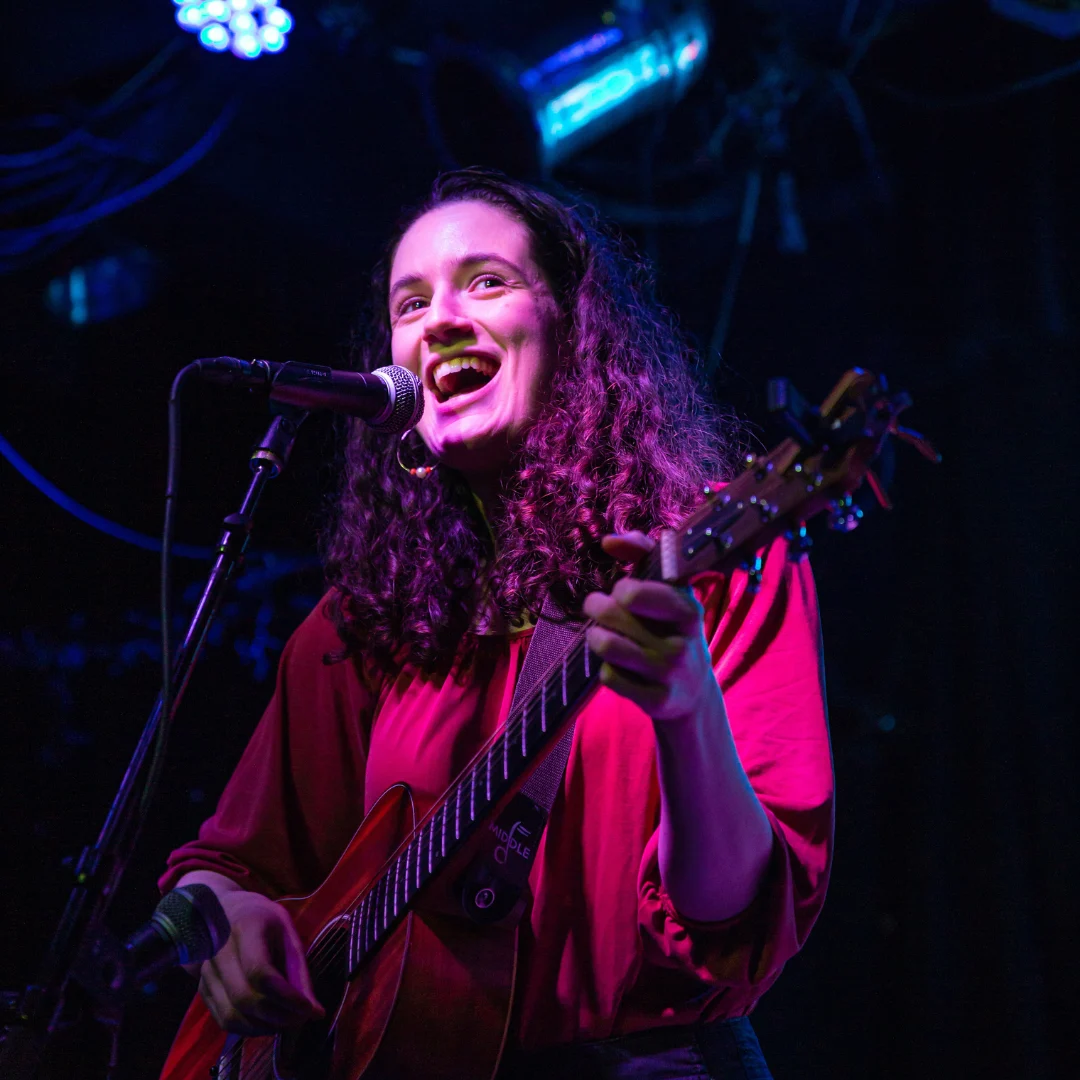 Sofía Campoamor performs live on stage with a ukulele, joyful and lit in blue and red stage light